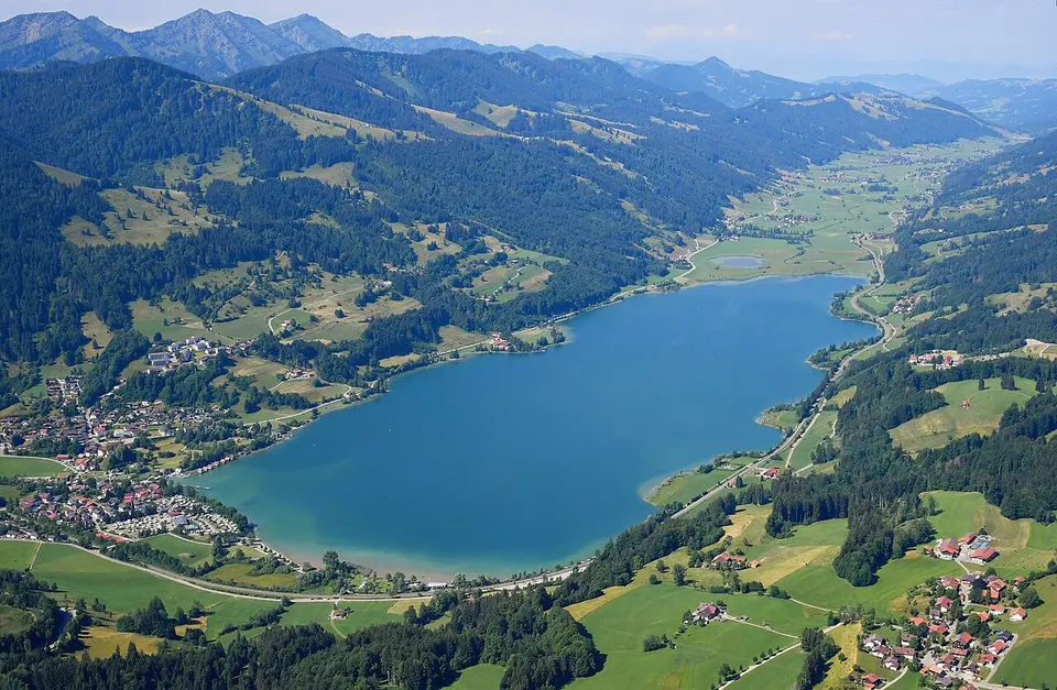 Luftaufnahme des Großen Alpsees im Allgäu mit grünen Hügeln und Bergen im Hintergrund